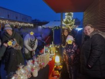 Mehrere Personen stehen warm gekleidet vorm Weihnachtsmarktstand. Im Hintergrund ist Beleuchtung